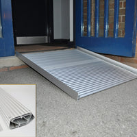 Rampa enrollable de aluminio para silla de ruedas de Able2 Shop, colocada frente a la puerta azul, con superficie acanalada antideslizante.